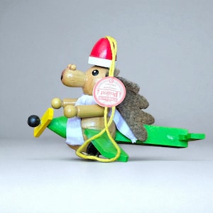 May include: A wooden hedgehog figurine wearing a red hat and a white scarf, riding a green airplane. The figurine has a brown body, a felt back, and a yellow string with a tag that reads "Steinbach Germany".