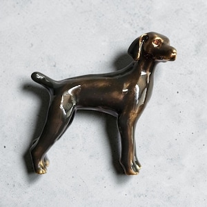May include: A bronze-colored dog figurine in a standing pose. The sculpture has a smooth, glossy finish and detailed features, including visible eyes and a curled tail. The dog is a dark brown color.