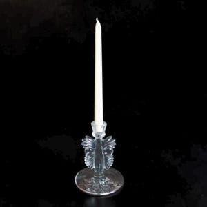 May include: A clear glass candlestick holder featuring a central stem with decorative wing-like protrusions and a circular base with an etched floral pattern, holding a single slender white taper candle against a solid black background.