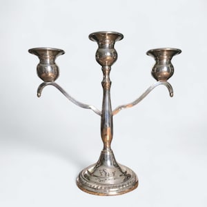 May include: A silver-colored candelabra with three candle holders. The candelabra has a central stem with two curved arms extending to the sides, each holding a candle cup. The base is round with decorative detailing.