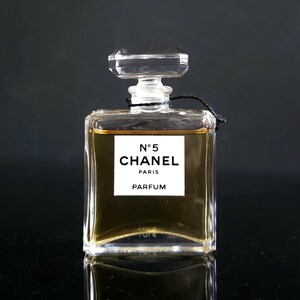 May include: A rectangular perfume bottle with a clear, faceted stopper. The bottle contains a golden-colored liquid and features a white label with the text "N&deg;5 CHANEL PARIS PARFUM". The bottle is set against a dark background.