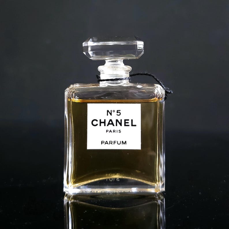 May include: A rectangular perfume bottle with a clear, faceted stopper. The bottle contains a golden-colored liquid and features a white label with the text "N&deg;5 CHANEL PARIS PARFUM". The bottle is set against a dark background.