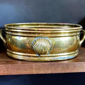 May include: A vintage brass planter with a seashell design. The rectangular planter has two curved handles and a decorative seashell in the center. The brass has a warm, aged patina, and the planter sits on a wooden shelf.