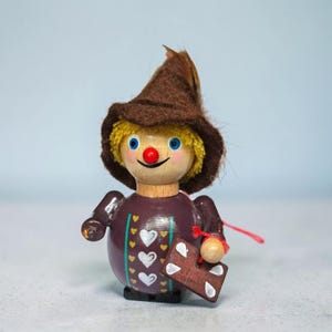 May include: A small wooden figurine wearing a brown felt hat and yellow hair. The round body is painted dark purple with white heart patterns and vertical green stripes. It holds a small brown rectangular sign with white heart details.