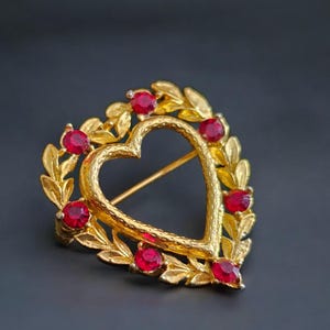 May include: A gold-toned heart-shaped brooch with a textured surface. The heart is surrounded by a laurel wreath design and adorned with red gemstones. The brooch has a pin closure on the back.
