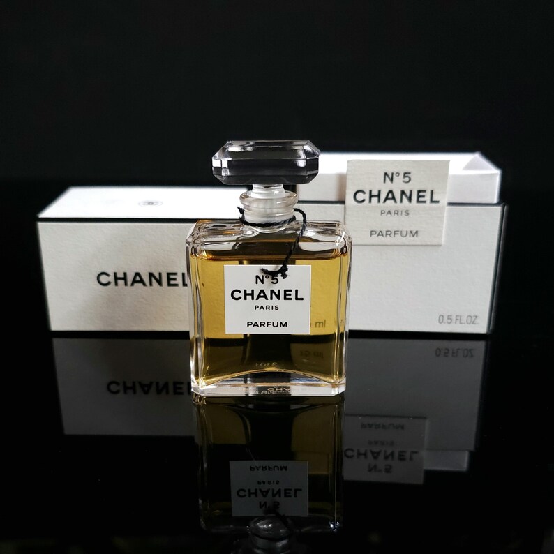 May include: A clear glass bottle of Chanel No. 5 Parfum with a black cap, a white label, and gold-colored liquid. The bottle is in front of a white box with the Chanel logo and text. The label reads "N&deg;5 CHANEL PARIS PARFUM".
