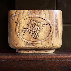 May include: A light brown, wooden container with a rectangular shape and a raised oval design featuring a cluster of grapes. The container has a textured, wood-grain appearance and sits on a small base.