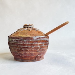May include: A handmade ceramic sugar bowl with a lid and a wooden spoon. The bowl is brown with horizontal ridges and a floral design on the lid. The spoon rests on the side of the bowl.