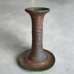 May include: A handcrafted, aged-looking candlestick holder with a flared top and a wide, circular base. The metal has a textured surface with a mix of red, green, and blue hues, creating a unique, antique appearance.