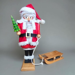 May include: A Santa Claus nutcracker wearing a red hat and coat with white faux fur trim, black belt, and glasses. The figure holds a green wooden Christmas tree in one hand and pulls a small wooden sled with a white cord attached to the waist.