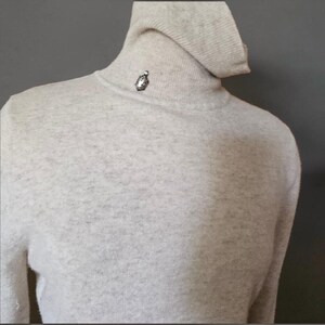 May include: A light beige roll-neck jumper with a soft, heathered texture. A small, silver-toned metallic emblem is attached to the centre of the neck, just below the collar fold, featuring a detailed, oval-shaped design with a dark centre.