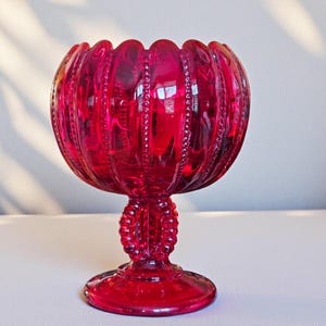 Könnte beinhalten: Ein rubinrotes Glas-Stielschale mit einem gewellten Rand und vertikalem Rippendesign. Die Schale hat einen runden Fuß und einen dekorativen Stiel mit einer Perlenstruktur. Das Glas reflektiert das Licht und hebt seine komplizierten Details hervor.