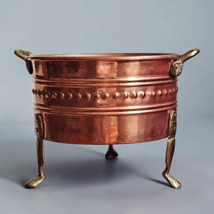May include: A round copper basin featuring a horizontal band of embossed circular studs around the center. The vessel includes two brass side handles and stands on three brass legs shaped like stylized feet, providing a stable base.