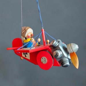May include: A hanging wooden toy aeroplane painted bright red with a pilot figure wearing a brown aviator cap. The plane features a silver engine block, a single orange propeller, and blue string rigging. A small white square with a black cross is visible.