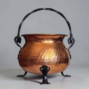 Peut inclure: Un chaudron rond en cuivre martelé doté d'une large ouverture et d'une finition métallique polie. Le récipient est soutenu par trois pieds métalliques sombres et incurvés et comprend une anse en métal torsadé assortie fixée aux boucles.