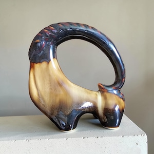 May include: A ceramic sculpture in the shape of a stylized animal, featuring a loop-shaped back. The piece is glazed in shades of brown, tan, and black, with a glossy finish. The sculpture is placed on a white surface.