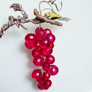 May include: A decorative cluster of faceted, ruby-red glass grapes hangs from a bronze-colored metal branch. The branch has a textured appearance and is adorned with a small, yellow and green leaf. The grapes are suspended against a white background.
