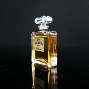 May include: A small, rectangular perfume bottle with a clear, faceted stopper. The bottle contains a golden liquid and features a label with the text "N&deg;5 CHANEL PARIS PARFUM". The bottle is reflected on a black surface.