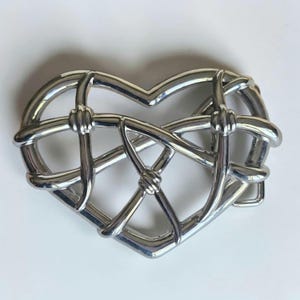 May include: A polished silver-tone metal belt buckle shaped like a heart. The design features a series of interconnected, smooth, rounded bars that form a cage-like structure within the heart outline, with small knotted details at the intersections.