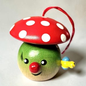 May include: A whimsical wooden mushroom ornament with a red cap featuring white polka dots. The mushroom has a green body, a red nose, and a smiling face. A yellow and blue bee is attached to the side, and a red string is attached to the top.