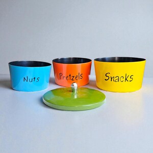 May include: Three colorful storage containers: blue with "Nuts", orange with "Pretzels", and yellow with "Snacks" in black text. A green lid with a silver knob sits in front. The containers have black interiors.