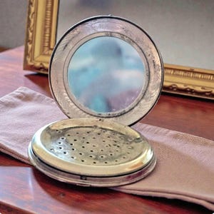 May include: An open, antique silver compact mirror with a round shape. The mirror is open, revealing a reflective surface. The bottom half of the compact has a perforated design. The compact sits on a mauve cloth, with a gold-framed mirror in the background.