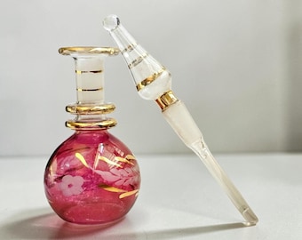 May include: A small, round, magenta glass perfume bottle with a clear glass neck and stopper. The bottle features gold-painted floral and leaf patterns, while the neck and stopper are accented with horizontal gold bands and frosted glass details.
