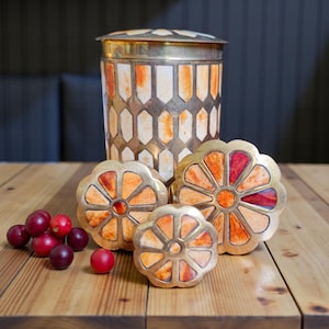 May include: A decorative set comprising a cylindrical container and three flower-shaped coasters. The container has a silver lid and a geometric patterned body. The coasters have a gold rim and orange and red stained glass designs.