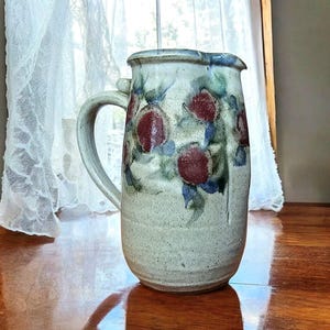 May include: A ceramic pitcher with a handle, featuring a cream-colored base adorned with red and blue floral designs. The pitcher is displayed on a wooden surface near a window with a sheer curtain.