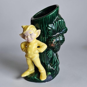 May include: A ceramic vase shaped like a dark green tree trunk with a textured bark surface and pinecone details. Attached to the front is a glossy yellow figure of a pointed-ear elf wearing a cap, standing with hands on hips and a smiling expression.