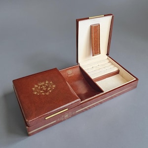 May include: A rectangular brown storage box with a hinged lid and a gold-colored emblem featuring a crown and floral design on the exterior. The interior contains a cream-colored velvet lining with slots for coins and the word COINS embossed.