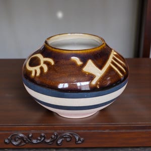 May include: A round ceramic vase with a wide opening. The vase is primarily brown with tan and white decorative elements. It features a blue stripe and a pink base. The vase is sitting on a dark wooden surface.
