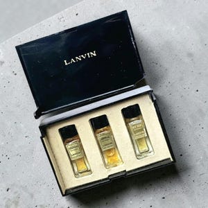 以下が含まれることがあります： 黒い箱に金色の文字で「LANVIN」と書かれています。中には3つのミニチュア香水ボトルが入っています。各ボトルには黒いキャップと、「SCANDAL」、「ARPEGE」、「MY SIN」という香りの名前が書かれたラベルが付いています。