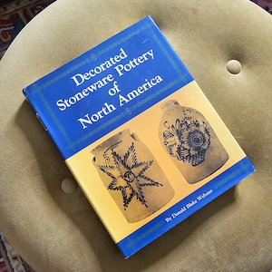 May include: A book titled "Decorated Stoneware Pottery of North America" with a blue cover and images of pottery. The book is open, displaying the title and illustrations of antique stoneware pieces.