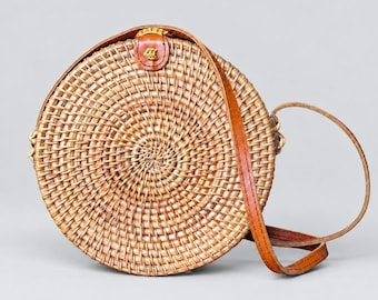 Op de afbeelding: Een ronde crossbodytas van gevlochten rotan met een natuurlijke lichtbruine afwerking. De tas heeft een cirkelvormig lichaam met een spiraalvormig vlechtpatroon, een bruine leren schouderband en een kleine bruine leren sluiting aan de bovenkant.