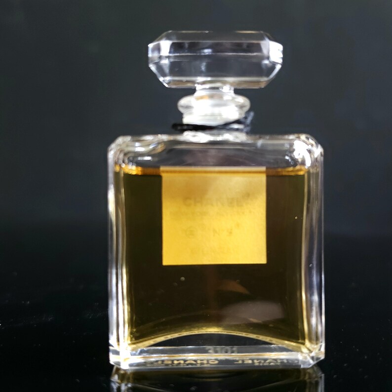 May include: A rectangular perfume bottle with a clear, faceted stopper. The bottle contains a golden liquid and has a gold label with the brand name and product number. The bottle is made of clear glass.