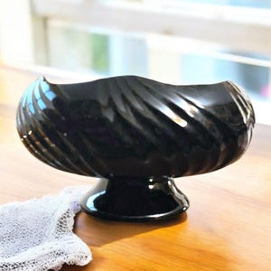 May include: A glossy black ceramic bowl with a pedestal base. The bowl has a wavy, swirled design and sits on a wooden surface. A white lace cloth is partially visible in the foreground. The bowl is likely for decorative or serving purposes.
