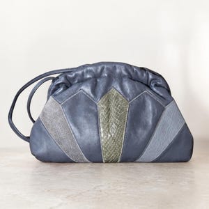 May include: A blue leather clutch bag with a drawstring closure and two handles. The front has a geometric design with grey and olive green panels. The bag is on a light-coloured surface.