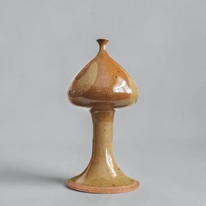 May include: A mushroom-shaped ceramic sculpture with a glossy finish. The cap features a mottled blend of light and dark brown tones, while the slender, flared stem is a lighter tan color with subtle speckles and a textured, unglazed base rim.