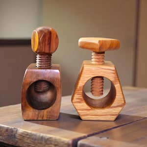 May include: Two wooden nutcrackers. One is a dark brown color with a rounded top, and the other is a lighter brown color with a flat top. Both have a screw mechanism and a hole for the nut.