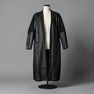 May include: A long, open-front black coat featuring a textured, crinkled finish. The garment has long sleeves, a straight hemline, and two large patch pockets on the front. The material appears to have a slight sheen, and the coat is displayed on a mannequin.
