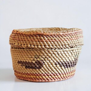 May include: Woven basket with a lid. The basket is made of natural fibers in shades of tan, brown, and peach. The lid fits snugly on top. The basket has a geometric pattern on the side.