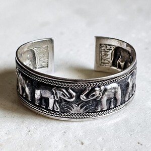 May include: Silver cuff bracelet with an elephant relief design. The bracelet has a braided border and rectangular ends with elephant imagery. The design is intricate and the metal has a polished finish.