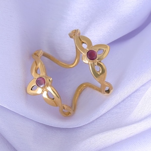 May include: A gold-coloured star-shaped ring featuring three flower-shaped designs, each with a small, round, red gemstone. The ring is set against a soft, light purple fabric.