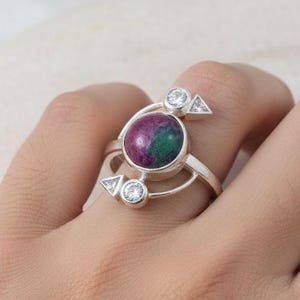 May include: A silver ring with a central gemstone in shades of red and green. The ring has a distinctive design, with two smaller clear gemstones and triangular details. The ring is displayed on a hand.