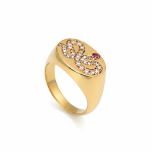 Peut inclure: Bague chevalière dorée ornée d'un motif de serpent. Le serpent est serti de petits pierres précieuses transparentes et d'un seul rubis rouge pour l'œil. La bague a une finition polie et un anneau arrondi.