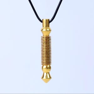 Puede incluir: Collar con colgante dorado con diseño cilíndrico. El colgante está suspendido de un cordón negro. La parte cilíndrica del colgante tiene una textura acanalada, con una base puntiaguda y una parte superior redondeada.