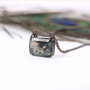 Puede incluir: Un collar con colgante rectangular con una piedra de ágata musgosa engastada en un marco de metal plateado. La piedra tiene un patrón nublado, verde y blanco. La cadena también es plateada.