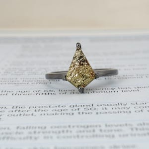 Peut inclure: Une bague géométrique avec un anneau sombre et une grande pierre triangulaire dorée. La pierre a une surface texturée avec des éclats bruns et dorés. La bague est posée sur un fond flou de texte imprimé.