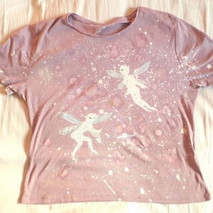 May include: A light purple short-sleeved t-shirt featuring a white paint splatter pattern across the front. The design includes two white fairy figures with wings, positioned centrally on the fabric, creating a layered and textured visual effect.
